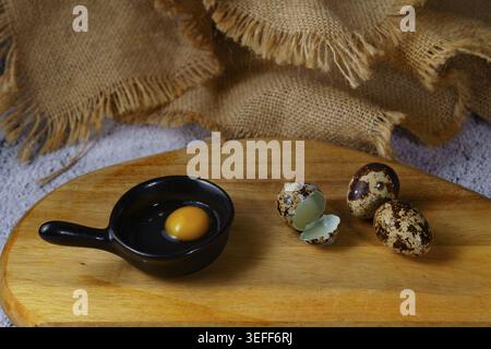 Quail egg yolk inside a small black ceramic bowl, with broken shells and whole quail eggs on a rustic wooden cutting board, preparing ingredients Stock Photo