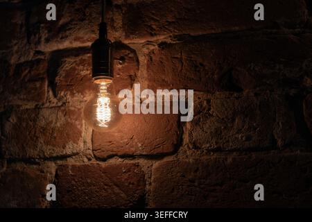 A vintage filament light bulb emits a warm amber glow as it hangs before a textured exposed brick wall background, creating a cozy, rustic atmosphere Stock Photo