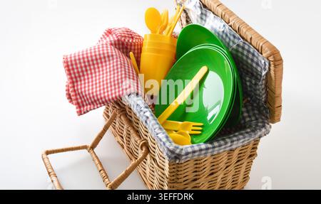 Open wicker picnic basket with colorful plastic plates, cups and cutlery on a clean white background, styled for outdoor dining and summer leisure. Stock Photo