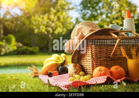 Wicker picnic basket with fruit, bread, wine and summer accessories on green grass in a sunny park, creating a warm outdoor leisure atmosphere. Stock Photo