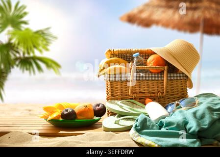 Beach picnic setup with basket, fruits, flip flops, sunglasses, sunscreen and hat on sand under sunny tropical light. Stock Photo