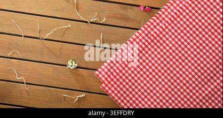 Top view of red gingham picnic fabric on a wooden table with small flowers and loose threads, creating a rustic summer picnic background. Stock Photo