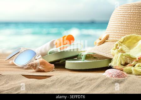Summer beach accessories with flip flops, sunglasses, straw hat and sunscreen on sandy shore with blurred ocean background and bright tropical sunligh Stock Photo