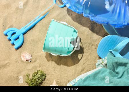 Colorful beach accessories on warm sand with flip flops, bucket, shovel, shells, and inflatable float in bright summer sunlight. Stock Photo