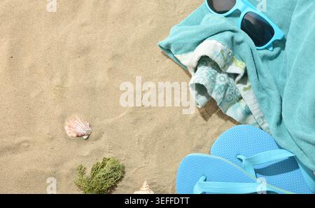 Beach essentials with blue flip flops, sunglasses, towels, seashells, and seaweed on soft sand under bright summer sunlight. Stock Photo