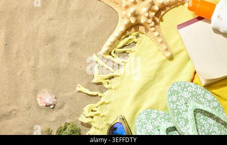 Sunny beach flat lay with starfish, flip flops, sunscreen, notebook, seashell, and yellow towel on textured sand background. Stock Photo