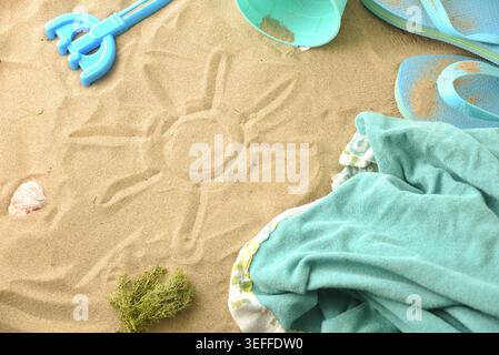 Hand-drawn sun symbol on sandy beach surrounded by summer accessories, flip flops, towel, shells, and bucket in bright daylight. Stock Photo
