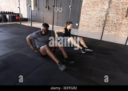 Diverse workout partners doing floor tricep dips on rubber mats near metal rig wearing gym shoes Stock Photo