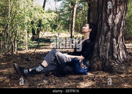 Young woman rests against large pine tree trunk in sunlit forest with eyes closed. Mental health awareness, emotional balance, inner peace concept Stock Photo