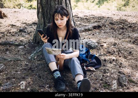 Person sits on ground using smartphone and paper map in forest. Hiking trail navigation, outdoor tourism activity, wilderness exploration trip. Stock Photo