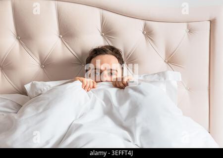 Frightened man lying in bed covering himself with blanket. Concept of fear, anxiety, insomnia and emotional stress. Stock Photo