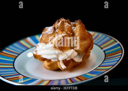 Delicious cream puff with whipped cream filling and powdered sugar on a colorful striped plate, isolated on black background. Stock Photo