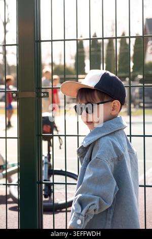 Cool boy in sunglasses and cap by fence on sunny playground Stock Photo