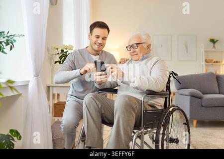 Smiling senior man in wheelchair using smartphone, exploring modern technology with caregiver. Stock Photo