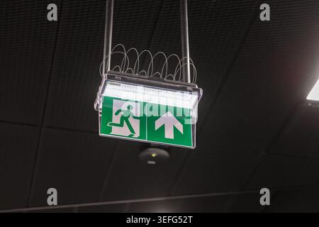 A bright glowing emergency exit sign with a directional arrow is mounted on a ceiling, representing safety regulations, clear navigation, disaster pre Stock Photo