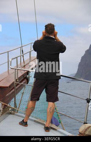 Man stands on boat deck, gazing at distant cliffs. He holds camera, capturing ocean's vastness. Sea winds stir his hair and shorts. Rugged coastline f Stock Photo