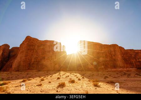 Sun burst creating dramatic light over towering eroded sandstone mountains and arid desert landscape with sparse vegetation in the Sharaan Nature Stock Photo