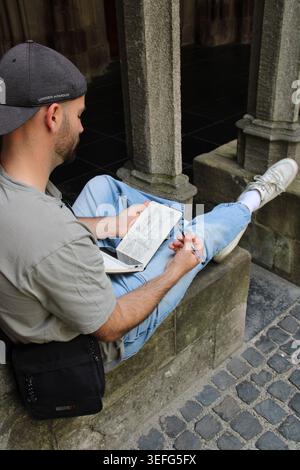 Man reads book, seated beneath stone archway. His posture is calm, focused on written text. Cobblestone floor and stone pillars frame the scene. Gentl Stock Photo