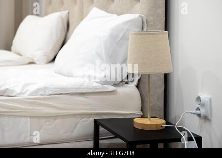 Close up of a modern wooden table lamp plugged into a white electrical socket beside a comfortable hotel bed, conveying minimalist home decor, interio Stock Photo