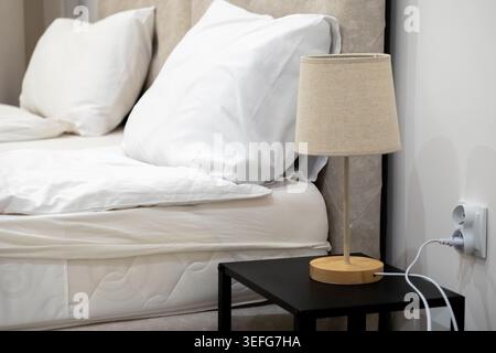Modern bedside table lamp on a dark side table with a plug connected to a wall socket near a comfortable bed, illustrating cozy home decor and interio Stock Photo