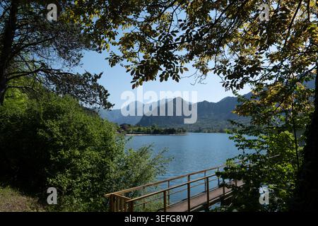 Leafy branches frame a scenic walkway overlooking the calm waters of Lake Idro and surrounding alpine mountains in a peaceful landscape of northern It Stock Photo