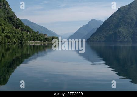 Calm reflective waters of Lake Idro mirror the surrounding alpine mountains beneath a soft blue sky in a tranquil natural landscape of northern Italy. Stock Photo