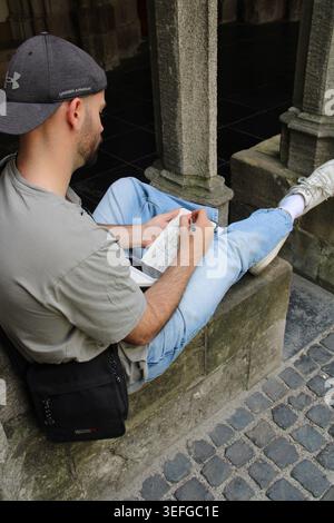 Man sits relaxed on stone step, reading paper. Wearing cap, casual jeans, and white sneakers. Sunlight highlights his focused, quiet expression. Histo Stock Photo