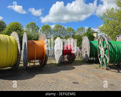 Large wooden reels loaded with colorful electrical cables are arranged in an outdoor industrial storage area under a bright blue sky with scattered cl Stock Photo