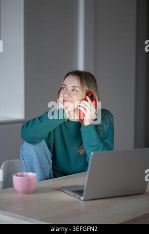 Pleased dreamy woman sits at table with laptop, coffee mug while talking on cellphone indoor. Thoughtful female looks away during phone conversation a Stock Photo
