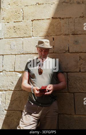 Casual traveler checking mobile phone in warm sunlight against vintage stone architecture during summer vacation in Europe. Stock Photo