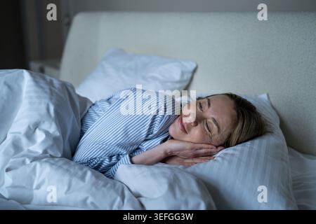 Happy serene mature woman lounging in cozy bed, awakening with smile, positive emotions, good mood. Stock Photo