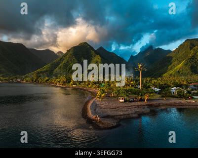 Aerial view of lush tropical mountains rising above a volcanic beach and turquoise lagoon under a dramatic sunset sky Moorea-Maiao, Windward Islands, Stock Photo