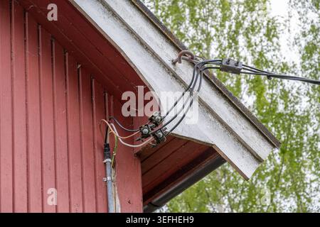 Close-up photograph of exposed electrical wires and cable connections mounted under the roof overhang of a traditional red wooden house. Stock Photo