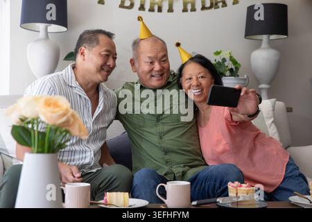 Asian family sitting closely on sofa, taking selfie with smartphone, displaying cake and party hats Stock Photo