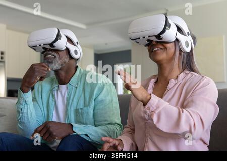 Senior couple wearing white VR headsets sitting on sofa in living room exploring virtual scene Stock Photo