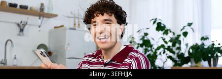 Young man with curly hair relaxes and smiles in his stylish apartment, banner Stock Photo