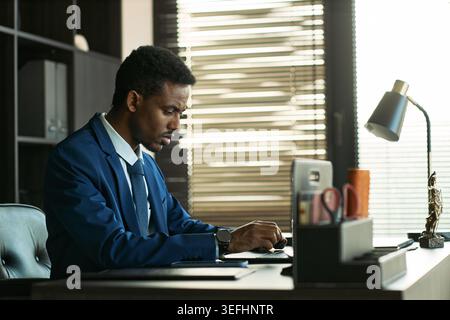 Medium shot of Black young adult businessman reviewing divorce case documents and typing at office desk, representing legal separation and family law services marketing Stock Photo