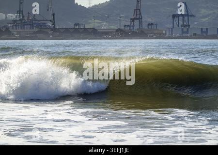 View of a powerful wave breaking at Ereaga beach with industrial port cranes and green hills in the background Algorta, Euskadi, Spain. Stock Photo