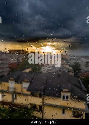 Raindrops cling to a window as dark storm clouds gather over Belgrade, Serbia, with blurred rooftops and golden evening light creating a dramatic urba Stock Photo