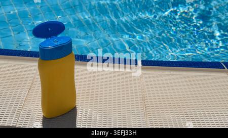 Yellow sunscreen bottle with blue cap placed on textured poolside near sparkling blue water of swimming pool under bright sunlight Stock Photo