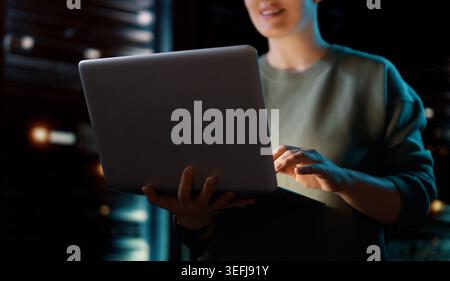 Confident professional woman working late on a laptop in a secure server room. The modern workspace highlights technology, innovation, and productivit Stock Photo