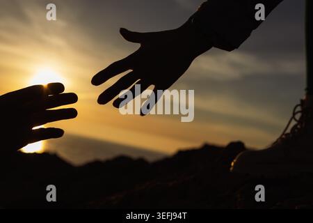 Silhouettes of two hands reaching across a glowing sunset, evoking support, rescue and human connection—offering hope, solidarity and compassion durin Stock Photo