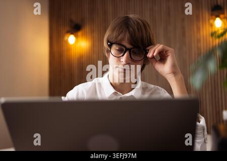 Young man wearing stylish glasses adjusting them while intently staring at a laptop screen, focusing on his work, studies, or online tasks in a modern Stock Photo