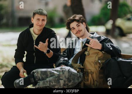 Two school friends relaxing on a bench with backpacks, smiling and making hand signs Stock Photo