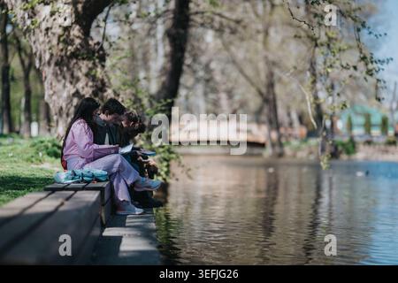 School friends sitting by a lake reading and studying together on a park bench Stock Photo