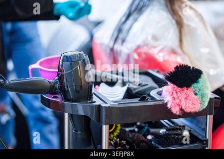 Professional hair stylist tools arranged on a salon trolley in beauty salon. Hair dryer, combs and brushes in hairstylist workspace with client in bac Stock Photo