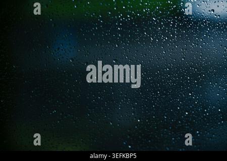 Raindrops on a window pane with blurry dark green trees and blue sky background, moody rainy weather abstract texture. Stock Photo