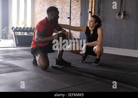 Kneeling man in red shirt holding bottle, squatting woman in black supporting in gym, battle ropes Stock Photo