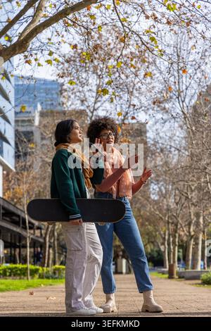 Diverse female friends walking on paved city path holding skateboard and smartphone wearing scarves Stock Photo