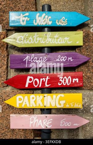 Stack of colorful hand painted wooden directional signs pointing to cafe distributeur plage port parking phare on weathered Breton stone wall coastal Stock Photo